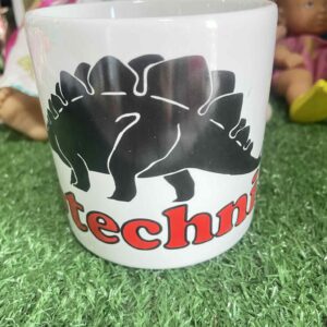 แก้วเซรามิกสีขาวที่มีรูปไดโนเสาร์สเตโกซอรัสสีดำและข้อความ “techni”